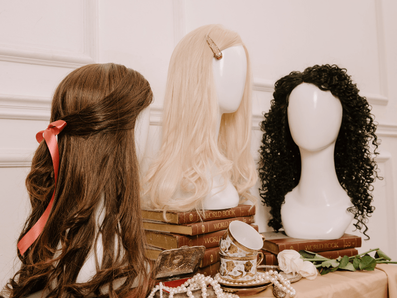 Three styled wigs displayed on mannequins with books, pearls, and vintage decor in the background