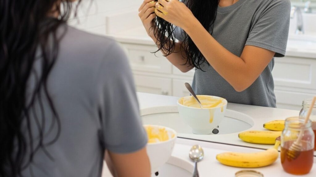 The image shows a banana and honey smoothing mask being applied evenly to damp hair, focusing on the lengths to reduce frizz and improve manageability. This DIY treatment supports softer, smoother hair when used as part of a balanced hair care routine, making it ideal for fine to medium hair types that need moisture without heaviness.